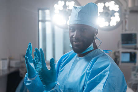 Smiling african american male surgeon with face mask in protective clothing in operating theatre....