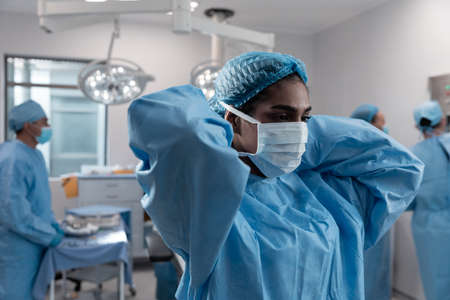 Mixed race female doctor putting on face mask standing in operating theatre. medicine, health and healthcare services during coronavirus covid 19 pandemic.の写真素材