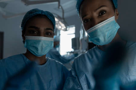 Diverse female doctors wearing face masks and surgical overalls looking at camera. medicine, health and healthcare services during coronavirus covid 19 pandemic.の写真素材
