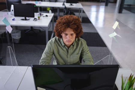 Mixed race businessman sitting in office in front of computer near sneeze shield. independent creative design business during covid 19 coronavirus pandemic.の写真素材