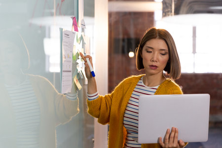 Asian businesswoman standing in front of whiteboard and writing holding laptop having video call....