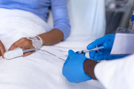 Midsection of african american male doctor giving iv injection to female patient in hospital bed. medicine, health and healthcare services during coronavirus covid 19 pandemic.の写真素材