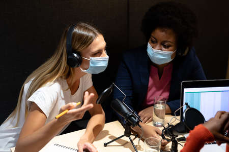 Diverse group of female business colleagues wearing masks in discussion speaking to microphones. recording podcast and business in a modern office during covid 19 coronavirus pandemic.の写真素材