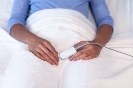 Midsection of mixed race female patient lying in hospital bed wearing fingertip pulse oximeter....