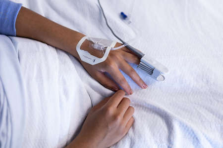 Midsection of mixed race female patient in hospital bed wearing fingertip pulse oximeter and iv tube. medicine, health and healthcare services.の写真素材