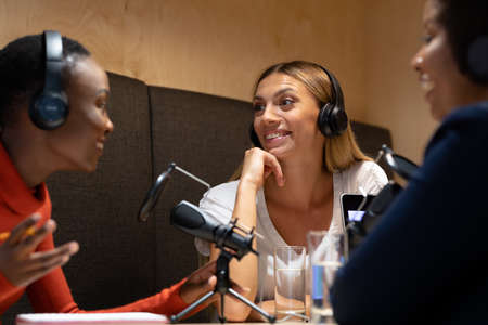 Diverse group of female business colleagues wearing headphones in discussion speaking to microphones. recording podcast and business in a modern office.の写真素材