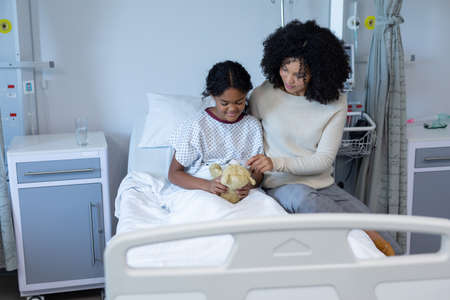 Mixed race mother comforting her sick daughter holding teddy bear in hospital bed. medicine, health and healthcare services.の写真素材