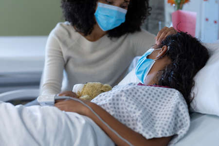 Mixed race mother and sick daughter in face masks in hospital, girl sleeping and holding teddy bear. medicine, health and healthcare services during covid 19 coronavirus pandemic.の写真素材