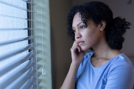 Mixed race female patient looking out of window in thought, resting head on hand. bored female...