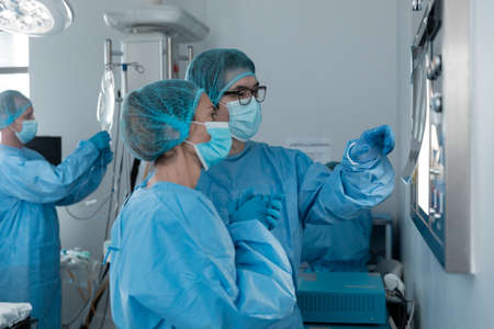 Diverse couple of male and female surgeons in operating theatre wearing face masks looking at screen. medicine, health and healthcare services during coronavirus covid 19 pandemic.の写真素材