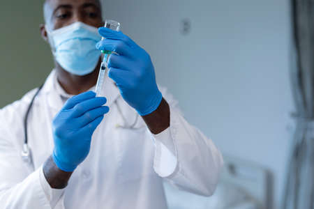 African american male doctor wearing face mask and gloves preparing covid vaccination. medicine, health and healthcare services during coronavirus covid 19 pandemic.の写真素材