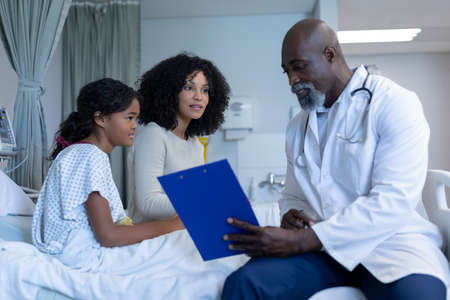 African american male doctor talking to mixed race mother and her sick daughter, in hospital bed. medicine, health and healthcare services.の写真素材