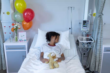 Sick mixed race girl asleep in hospital bed wearing fingertip pulse oximeter and holding teddy bear. medicine, health and healthcare services.の写真素材