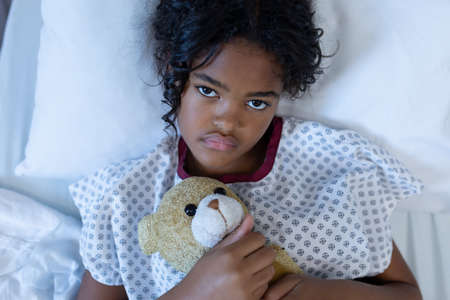 Portrait of sick mixed race girl lying in hospital bed holding teddy bear. medicine, health and...