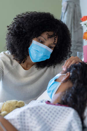 Mixed race mother and sick daughter in face masks in hospital, girl holding teddy bear. medicine,...