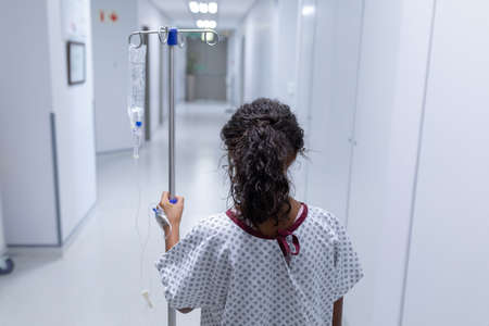 Rear view of sick mixed race girl walking in hospital corridor with iv drip bag on stand. medicine,...