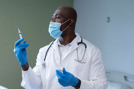 African american male doctor wearing face mask and gloves preparing covid vaccination in hospital. medicine, health and healthcare services during coronavirus covid 19 pandemic.の写真素材
