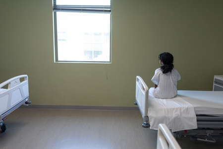 Rear view of sick mixed race girl sitting on hospital bed looking out of window. medicine, health and healthcare services.の写真素材