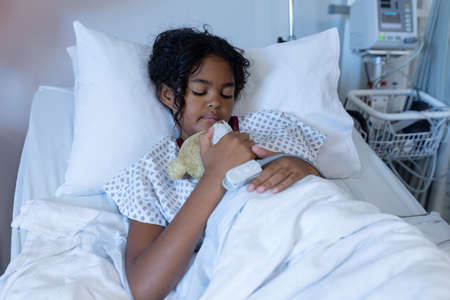 Mixed race sick girl asleep in hospital bed wearing fingertip pulse oximeter and holding teddy bear. medicine, health and healthcare services.の写真素材