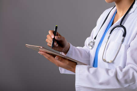 Mid section of african american female doctor using graphic tablet against grey background. healthcare and medical professionalism conceptの写真素材