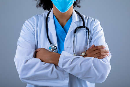 Mid section of female doctor wearing face mask standing with arms crossed against grey background. healthcare and medical professionalism conceptの写真素材