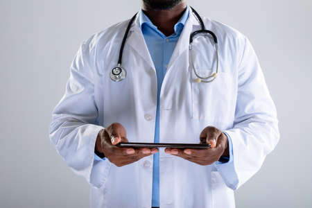 Mid section of african american male doctor holding digital tablet against grey background. healthcare and medical professionalism conceptの写真素材