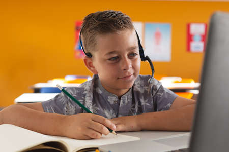 Caucasian boy sitting at desk in classroom wearing headset, looking at laptop and smiling....