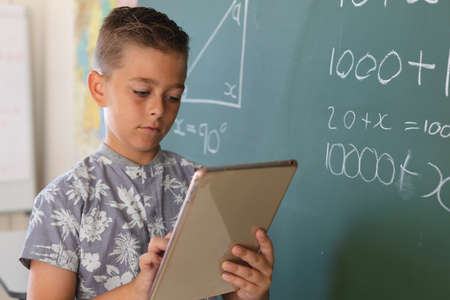Caucasian boy standing at chalkboard in classroom and using tablet during maths lesson. childhood and education at elementary school.の写真素材