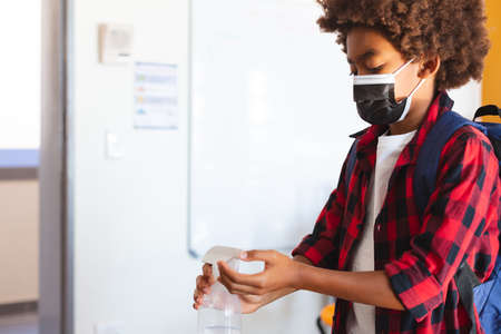 African american schoolboy in face mask standing in classroom disinfecting hands. childhood and education at elementary school during coronavirus covid19 pandemic.の写真素材