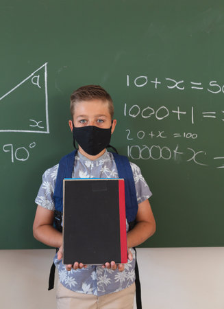 Portrait of caucasian boy in face mask standing at chalkboard with geometry on it holding books. childhood and education at elementary school during coronavirus covid19 pandemic.の写真素材