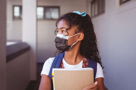 African american schoolgirl in face mask standing in school corridor holding books. childhood and...