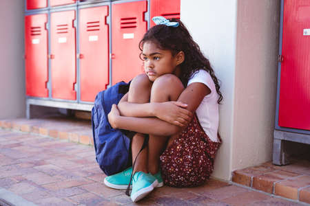 Unhappy african american schoolgirl sitting by lockers in school corridor with schoolbag. childhood and education at elementary school.の写真素材