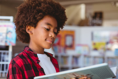 Smiling african american schoolboy reading book standing in school library. childhood and education...