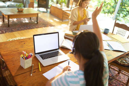 Asian girl using laptop with blank screen, raising hand, learning online her mum in background. at home in isolation during quarantine lockdown.の写真素材