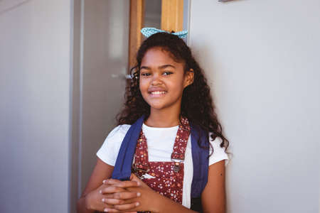 Portrait of smiling african american schoolgirl standing in school corridor wearing schoolbag....