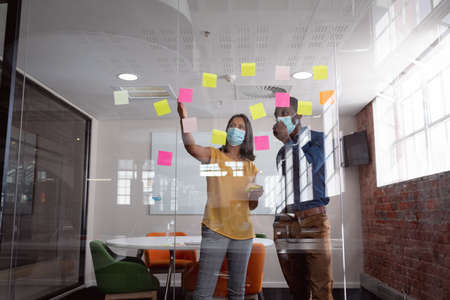 Diverse male and female colleague in face masks brainstorming, reading memo notes on glass wall....