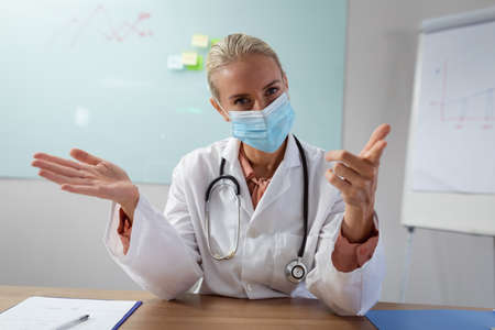 Caucasian female doctor wearing face mask sitting at desk in office gesturing during video call.