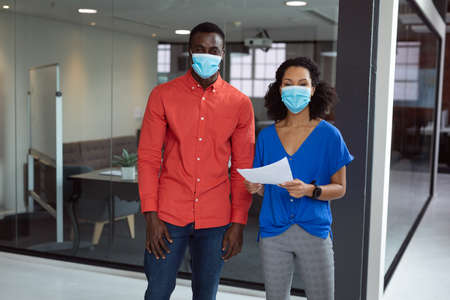 Portrait of diverse male and female colleague in face masks holding paperwork in office.