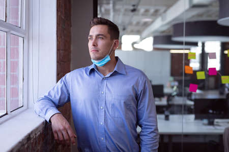 Portrait of thoughtful caucasian businessman in lowered face mask looking out of window.