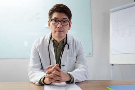 Asian male doctor sitting at desk in office listening during video call.の写真素材