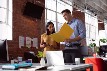 Serious caucasian male and female colleague standing at desk discussing tablet and folder. working...