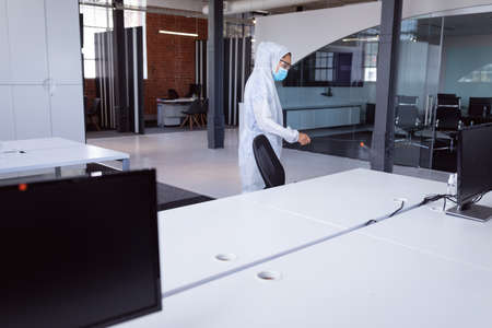 Cleaner wearing ppe suit, glasses and mask disinfecting office workspace.