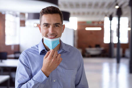 Portrait of happy caucasian businessman lowering face mask and smiling to camera standing in office. working in business at a modern office during pandemic.の写真素材
