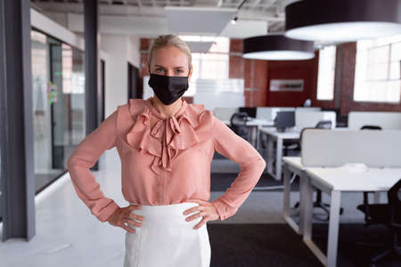 Portrait of caucasian businesswoman in face mask standing in office with hands on hips. working in business at a modern office during pandemic.の写真素材