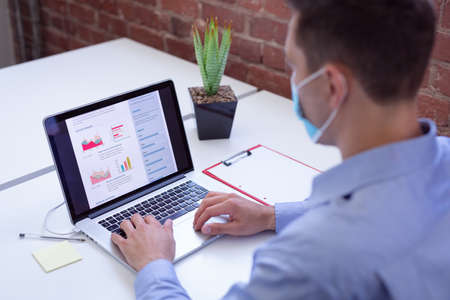 Caucasian businessman wearing face mask sitting at desk in office using laptop. working in business...