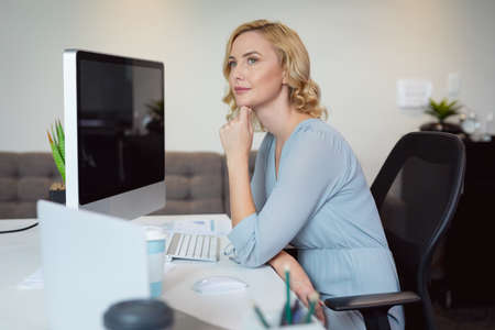 Thoughtful caucasian businesswoman sitting at desk using computer, looking away in thought. working in business at a modern office.の写真素材