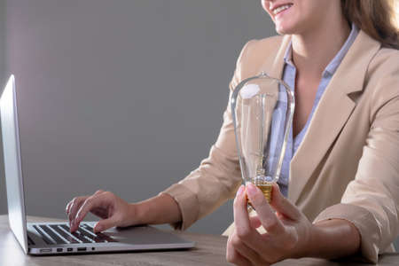 Smiling caucasian businesswoman holding light bulb using laptop, isolated on grey background. business, technology, communication and growth concept.の写真素材