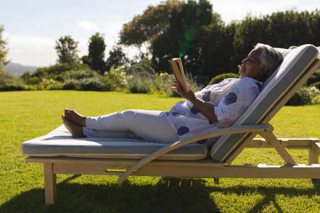 Senior african american woman reading book in deckchair in sunny garden. retreat, retirement and...