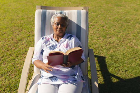 Senior african american woman reading book in deckchair in sunny garden. retreat, retirement and happy senior lifestyle concept.の写真素材