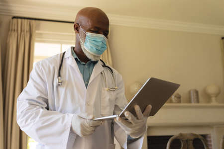 Portrait of senior african american male doctor in face mask and lab coat using laptop. retirement and senior lifestyle during covid 19 pandemic concept.の写真素材